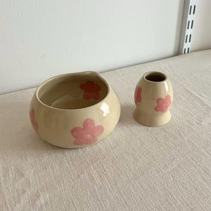 Pu&ograve; includere: Una ciotola in ceramica color crema e un piccolo vaso abbinato, entrambi caratterizzati da un motivo dipinto a mano di fiori a cinque petali in rosa tenue e rosso scuro. La ciotola ha una forma arrotondata con una leggera rientranza sul bordo.