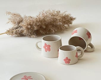 May include: Three white ceramic mugs with pink flower designs. The mugs have a rounded shape and a handle on the side.