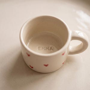 May include: A small, off-white ceramic mug featuring a smooth, rounded handle and a series of tiny, hand-painted red hearts scattered across the exterior. The interior base of the mug is clearly stamped with the name EMMA in capital letters.