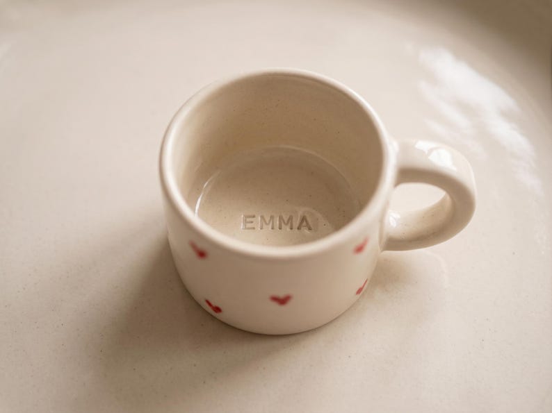 May include: A small, off-white ceramic mug featuring a smooth, rounded handle and a series of tiny, hand-painted red hearts scattered across the exterior. The interior base of the mug is clearly stamped with the name EMMA in capital letters.