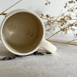 May include: A cream-colored ceramic mug with a smooth finish and a handle on the right side. The interior base of the mug features the stamped date 08.17.23 in a simple, dark font. The mug is viewed from directly above, showing the circular opening.