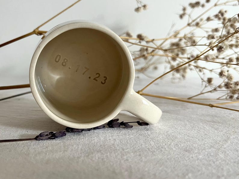 May include: A cream-colored ceramic mug with a smooth finish and a handle on the right side. The interior base of the mug features the stamped date 08.17.23 in a simple, dark font. The mug is viewed from directly above, showing the circular opening.