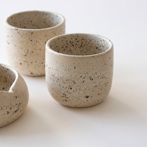May include: Three speckled ceramic cups and a small bowl are arranged on a white surface. The cups are cylindrical with rounded bottoms. The bowl has a curved, asymmetrical shape. The ceramic pieces have a neutral, speckled color.