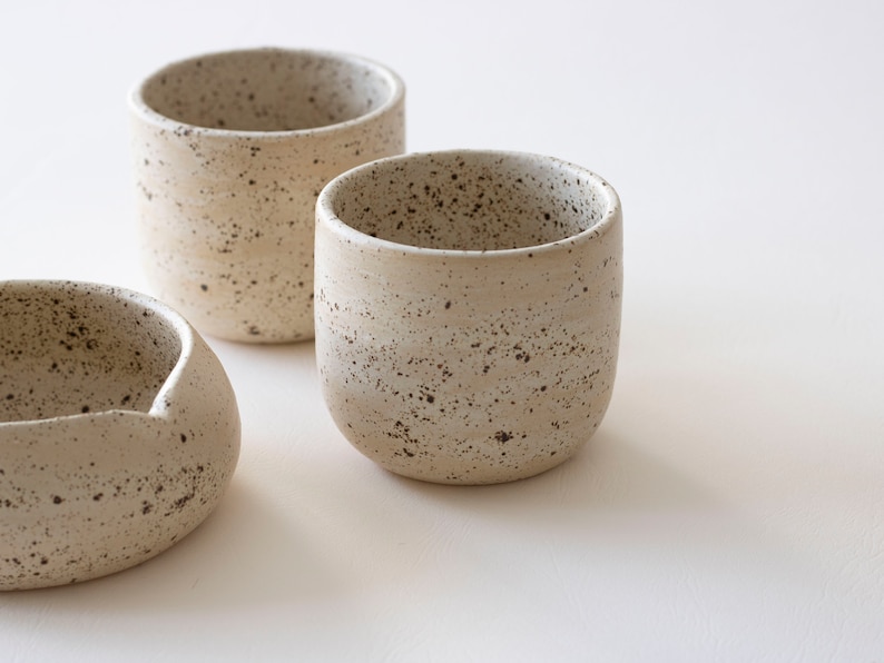 May include: Three speckled ceramic cups and a small bowl are arranged on a white surface. The cups are cylindrical with rounded bottoms. The bowl has a curved, asymmetrical shape. The ceramic pieces have a neutral, speckled color.