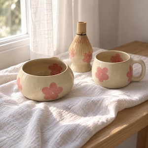 Pu&ograve; includere: Una ciotola in ceramica color crema con beccuccio, una tazza abbinata con manico e un frustino per matcha in bamb&ugrave;, tutti decorati con motivi floreali rosa dipinti a mano. Gli oggetti sono disposti insieme, mostrando un design floreale coerente.