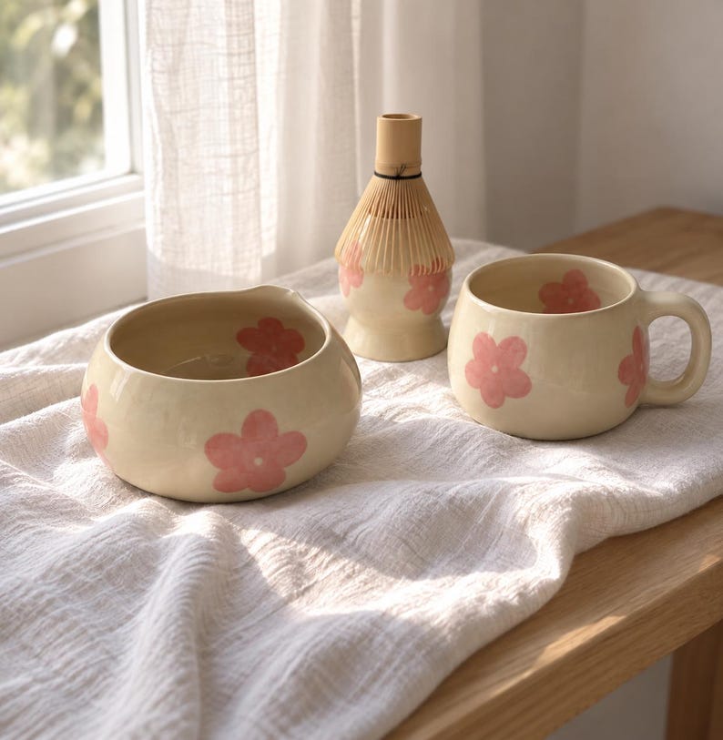 Pu&ograve; includere: Una ciotola in ceramica color crema con beccuccio, una tazza abbinata con manico e un frustino per matcha in bamb&ugrave;, tutti decorati con motivi floreali rosa dipinti a mano. Gli oggetti sono disposti insieme, mostrando un design floreale coerente.