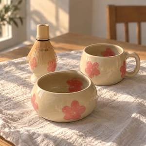Pu&ograve; includere: Un set da matcha in ceramica composto da una tazza color crema, una piccola ciotola e un frustino in bamb&ugrave;. Ogni pezzo &egrave; decorato con motivi floreali rosa dipinti a mano. La tazza ha un corpo arrotondato con manico, la ciotola ha un bordo liscio.