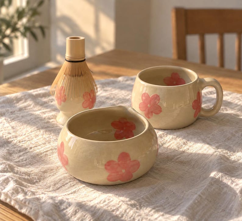 Pu&ograve; includere: Un set da matcha in ceramica composto da una tazza color crema, una piccola ciotola e un frustino in bamb&ugrave;. Ogni pezzo &egrave; decorato con motivi floreali rosa dipinti a mano. La tazza ha un corpo arrotondato con manico, la ciotola ha un bordo liscio.