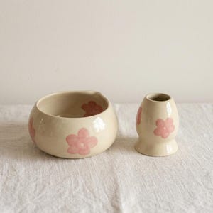 Pu&ograve; includere: Una ciotola in ceramica color crema e un piccolo vaso abbinato, entrambi decorati con motivi floreali rosa chiaro dipinti a mano. La ciotola ha una forma arrotondata con un leggero beccuccio, mentre il vaso presenta un collo stretto e una base svasata.