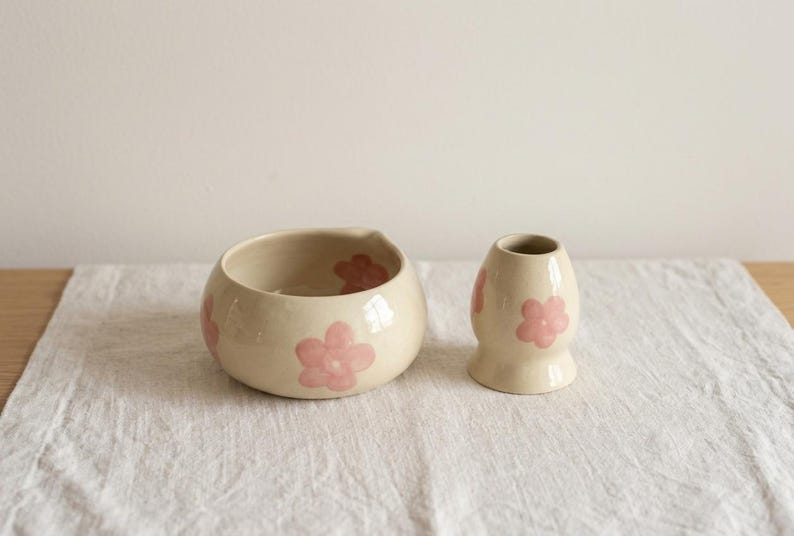 Pu&ograve; includere: Una ciotola in ceramica color crema e un piccolo vaso abbinato, entrambi decorati con motivi floreali rosa chiaro dipinti a mano. La ciotola ha una forma arrotondata con un leggero beccuccio, mentre il vaso presenta un collo stretto e una base svasata.