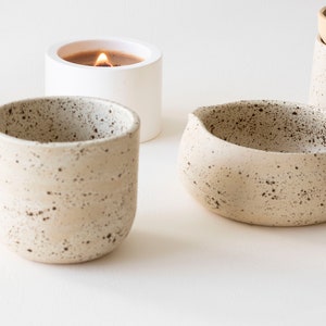 May include: A collection of ceramic home decor items. Includes a speckled beige candle holder with a lit candle, a small speckled beige bowl, and a speckled beige cup. The items are arranged on a white surface.