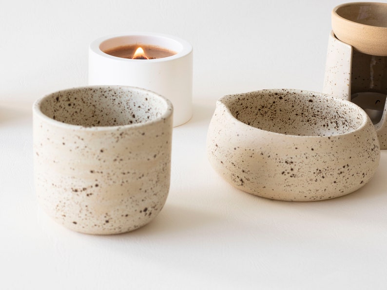 May include: A collection of ceramic home decor items. Includes a speckled beige candle holder with a lit candle, a small speckled beige bowl, and a speckled beige cup. The items are arranged on a white surface.