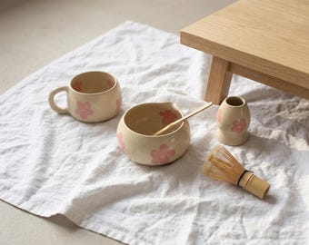 May include: A ceramic matcha set featuring a cream-colored mug, a bowl with a pouring spout, and a whisk holder, all decorated with pink flower motifs. A bamboo whisk and a bamboo scoop are included to complete this tea preparation collection.