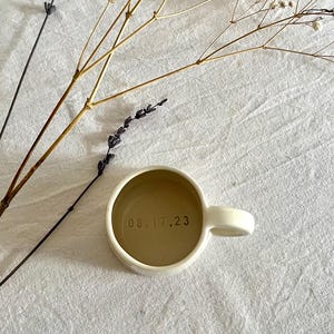 May include: A cream-colored ceramic mug with a rounded handle and a smooth finish. The interior base of the mug features the date 08.17.23 stamped in dark, clear lettering. The mug is viewed from directly above, showcasing its circular shape.