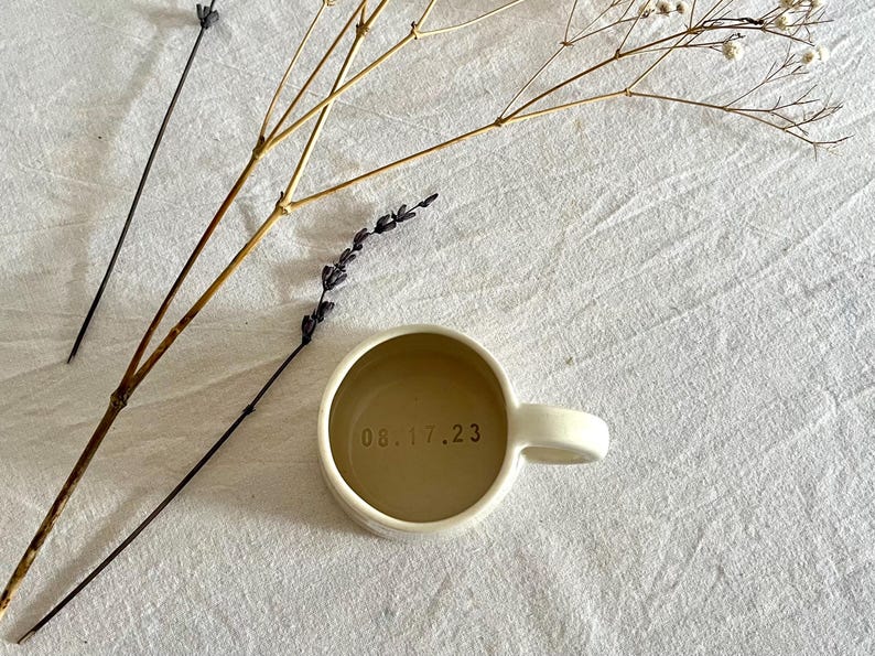 May include: A cream-colored ceramic mug with a rounded handle and a smooth finish. The interior base of the mug features the date 08.17.23 stamped in dark, clear lettering. The mug is viewed from directly above, showcasing its circular shape.