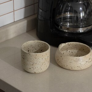 May include: Two speckled ceramic pieces: a cylindrical cup and a small, rounded bowl. The off-white pottery has dark brown speckles. A black coffee maker is in the background, suggesting a kitchen setting.