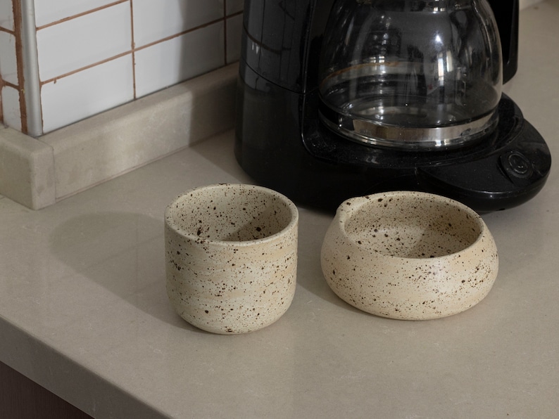 May include: Two speckled ceramic pieces: a cylindrical cup and a small, rounded bowl. The off-white pottery has dark brown speckles. A black coffee maker is in the background, suggesting a kitchen setting.