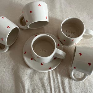 May include: A set of four white ceramic mugs and one matching saucer, each decorated with small red heart patterns. One mug features the text LOVE YOU inside, while the central mug on the saucer displays the text I&H embossed on the interior base.