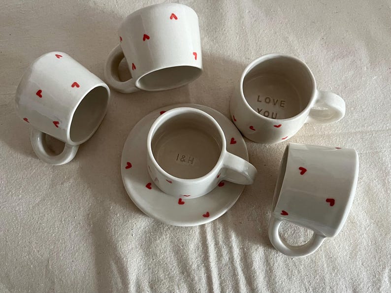 May include: A set of four white ceramic mugs and one matching saucer, each decorated with small red heart patterns. One mug features the text LOVE YOU inside, while the central mug on the saucer displays the text I&H embossed on the interior base.