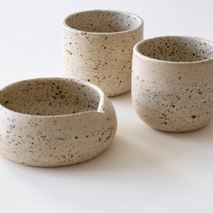 May include: Three speckled ceramic pieces: a small bowl with a spout and two cylindrical cups. The off-white pottery has dark brown speckles, creating a rustic, handcrafted aesthetic. The pieces are arranged on a white surface.