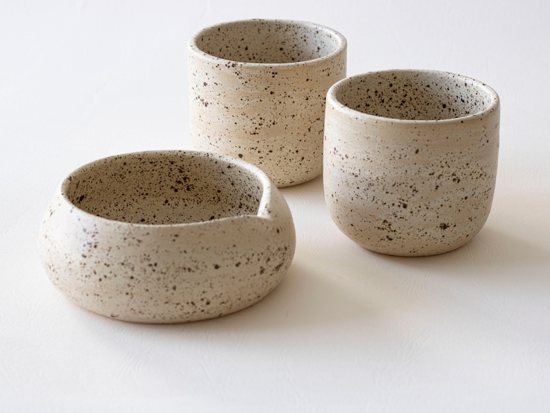 May include: Three speckled ceramic pieces: a small bowl with a spout and two cylindrical cups. The off-white pottery has dark brown speckles, creating a rustic, handcrafted aesthetic. The pieces are arranged on a white surface.
