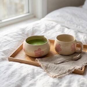Pu&ograve; includere: Una ciotola in ceramica riempita di t&egrave; matcha verde e una tazza in ceramica abbinata, entrambe caratterizzate da un motivo a fiori rosa chiaro. Questi oggetti poggiano su un vassoio rettangolare in legno accanto a un cucchiaino d'argento su un tovagliolo di lino beige.