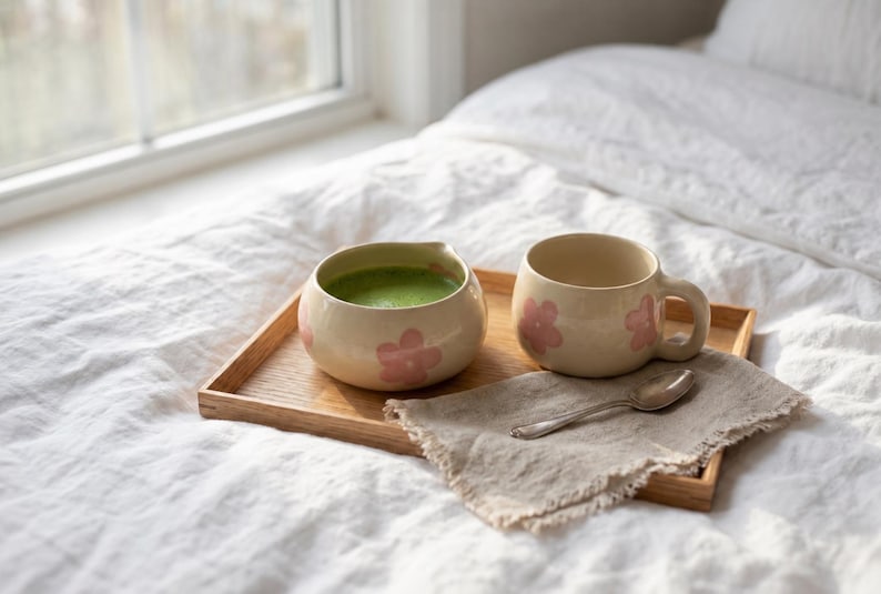 Pu&ograve; includere: Una ciotola in ceramica riempita di t&egrave; matcha verde e una tazza in ceramica abbinata, entrambe caratterizzate da un motivo a fiori rosa chiaro. Questi oggetti poggiano su un vassoio rettangolare in legno accanto a un cucchiaino d'argento su un tovagliolo di lino beige.