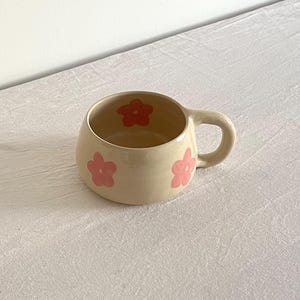 Könnte beinhalten: Eine beige Keramiktasse mit abgerundeter Form und einem Henkel. Die Tasse hat ein rosafarbenes Blumenmuster.