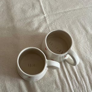 May include: Two off-white ceramic mugs with small red heart patterns on the exterior. One mug features the text I&H printed on the interior base, while the other mug displays the date 08.17.23 printed on its interior base. Both mugs have handles.