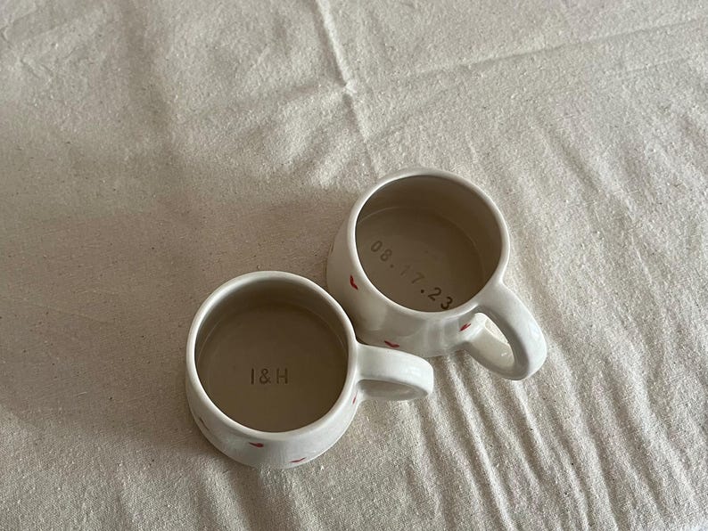 May include: Two off-white ceramic mugs with small red heart patterns on the exterior. One mug features the text I&H printed on the interior base, while the other mug displays the date 08.17.23 printed on its interior base. Both mugs have handles.