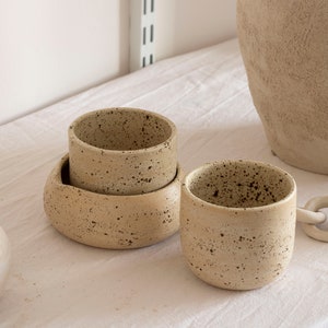 May include: A collection of speckled ceramic pottery in neutral tones. The set includes a small cup, a bowl, and a larger vessel, all with a textured surface. The pieces are arranged on a white surface, suggesting a minimalist aesthetic.