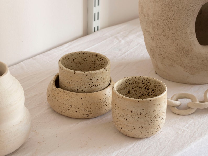 May include: A collection of speckled ceramic pottery in neutral tones. The set includes a small cup, a bowl, and a larger vessel, all with a textured surface. The pieces are arranged on a white surface, suggesting a minimalist aesthetic.