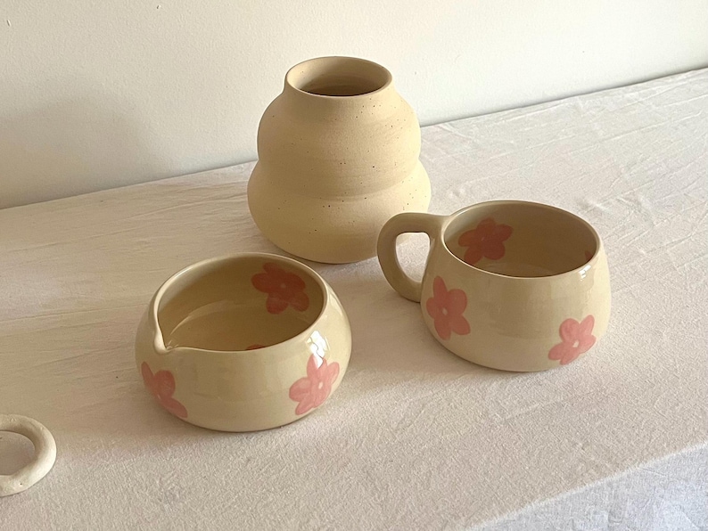 Pu&ograve; includere: Un set di tre pezzi in ceramica composto da un vaso beige opaco dalla silhouette arrotondata, una piccola ciotola con beccuccio e una tazza. Sia la ciotola che la tazza presentano una finitura color crema lucida decorata con fiori rosa dipinti.