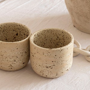May include: Two beige ceramic mugs with a speckled pattern. The mugs have a rounded shape and a smooth, matte finish. A white chain-like decorative element is visible in the background, along with a portion of a larger beige ceramic vessel.