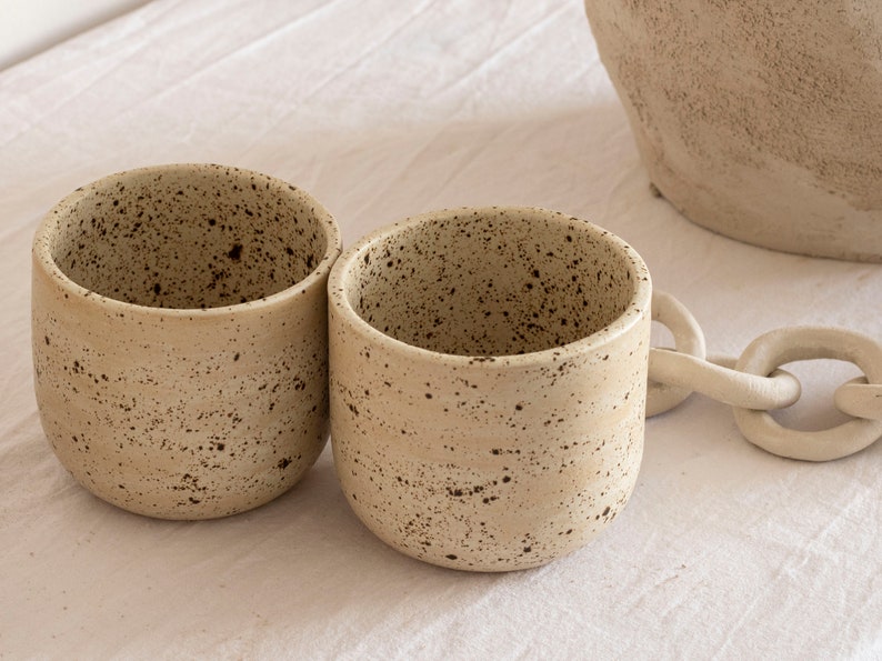 May include: Two beige ceramic mugs with a speckled pattern. The mugs have a rounded shape and a smooth, matte finish. A white chain-like decorative element is visible in the background, along with a portion of a larger beige ceramic vessel.