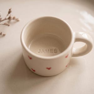 May include: A small, off-white ceramic mug featuring a rounded handle and a pattern of tiny red hearts on the exterior. The interior base of the mug is embossed with the name JAMES in capital letters, centered within the bottom of the vessel.