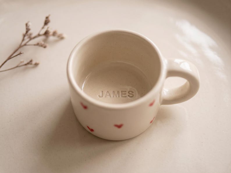 May include: A small, off-white ceramic mug featuring a rounded handle and a pattern of tiny red hearts on the exterior. The interior base of the mug is embossed with the name JAMES in capital letters, centered within the bottom of the vessel.