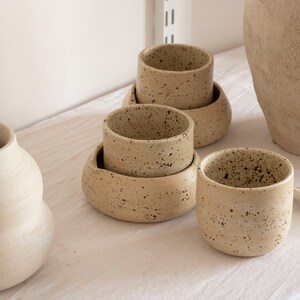 May include: A collection of handmade ceramic cups and saucers in a neutral beige color with dark speckles. The cups are cylindrical, and some are stacked on rounded saucers. Other ceramic pieces are in the background.