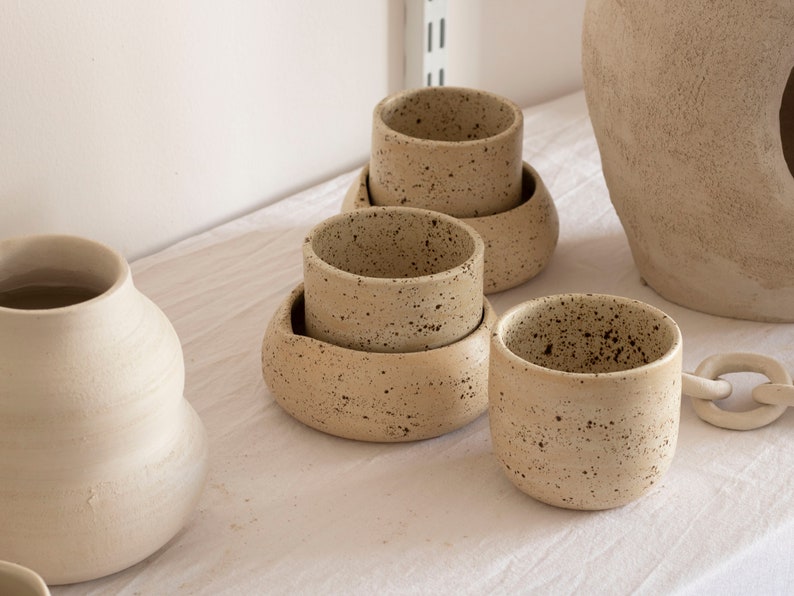 May include: A collection of handmade ceramic cups and saucers in a neutral beige color with dark speckles. The cups are cylindrical, and some are stacked on rounded saucers. Other ceramic pieces are in the background.