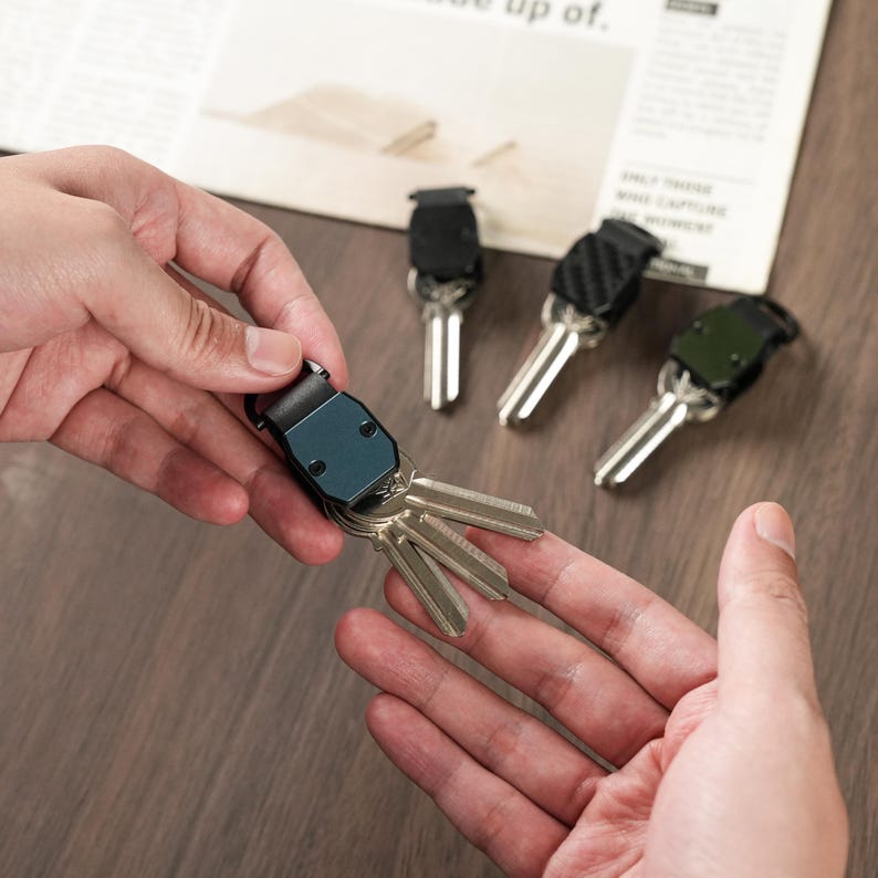 May include: A blue metal key organizer holding three silver keys, with three additional key organizers in black and carbon fiber patterns displayed nearby. The organizers feature a compact, folding design with visible screws and a secure attachment loop.