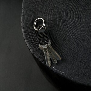 May include: A black carbon fiber key organizer featuring a silver metal carabiner ring and a central screw mechanism. The device holds a set of three metallic keys in a compact arrangement, showcasing a woven texture on the main rectangular body.