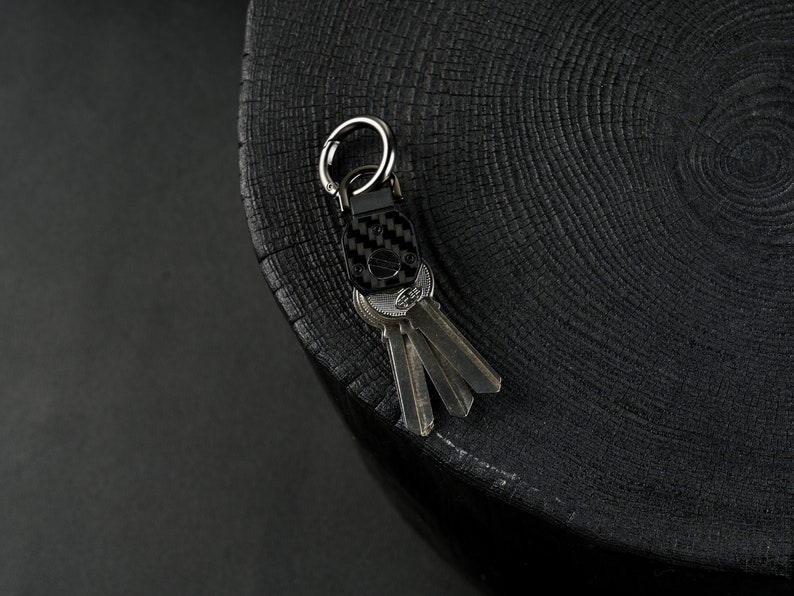 May include: A black carbon fiber key organizer featuring a silver metal carabiner ring and a central screw mechanism. The device holds a set of three metallic keys in a compact arrangement, showcasing a woven texture on the main rectangular body.