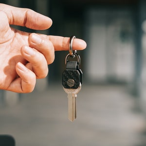 以下が含まれることがあります： 銀色の鍵に、黒いカーボンファイバー製のキーホルダーが銀色のリングで取り付けられています。鍵は指で持たれています。