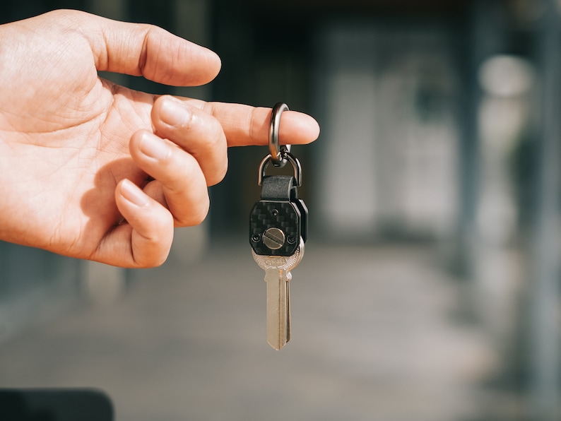May include: A metal key attached to a dark, carbon fiber-patterned key fob with a silver-toned screw. The key fob is connected to a circular metal ring, featuring a sleek, modern design with a polished finish on the key blade and a matte texture.