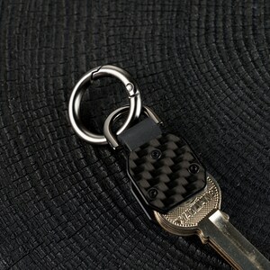 May include: A metal key organizer featuring a carbon fiber textured plate secured with two black screws. The device includes a circular gunmetal carabiner ring attached to a black connector, with a metallic key partially inserted into the base.