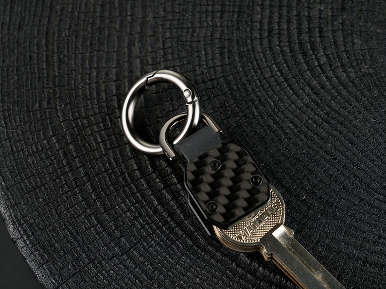 May include: A metal key organizer featuring a carbon fiber textured plate secured with two black screws. The device includes a circular gunmetal carabiner ring attached to a black connector, with a metallic key partially inserted into the base.