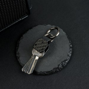 May include: A metal keychain featuring a carbon fiber patterned top section with four small screws, attached to a dark gunmetal ring. The keychain holds a silver-colored key with the word "YIOLANG" embossed on the head of the key blade.