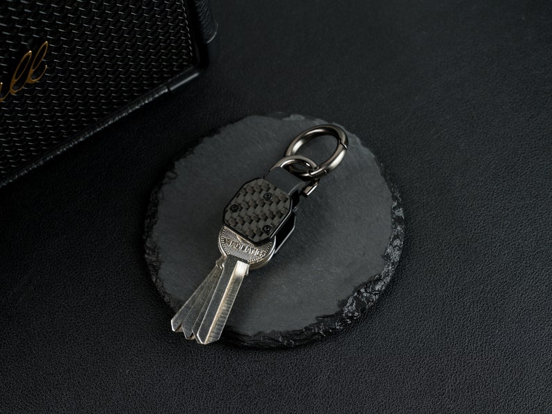 May include: A metal keychain featuring a carbon fiber patterned top section with four small screws, attached to a dark gunmetal ring. The keychain holds a silver-colored key with the word "YIOLANG" embossed on the head of the key blade.