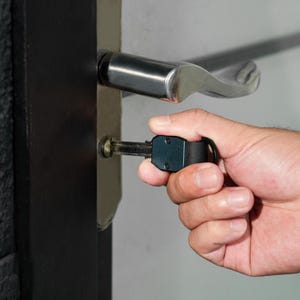 May include: A dark metallic key with a rectangular head and a black circular ring attachment inserted into a silver door lock cylinder. The lock is mounted on a black door frame featuring a horizontal silver lever handle above the keyhole.