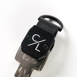 May include: A black metal key organizer featuring a matte finish and a white logo with the letters C and L separated by a diagonal line. The device is secured with two small screws and includes a circular ring attachment holding a silver ISEO key.